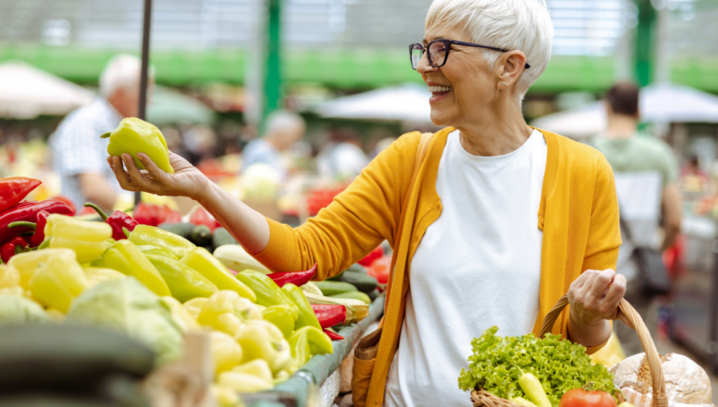Eine Dame mit Brille und Korb kauft auf dem Wochenmarkt frisches Gemüse ein.