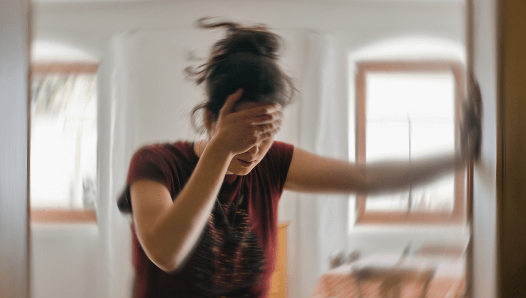Schlaganfall-Symptome bei Frauen: Eine junge Frau greift sich an den Kopf, ihre Umgebung ist verschwommen, um einen Schwindelanfall anzudeuten