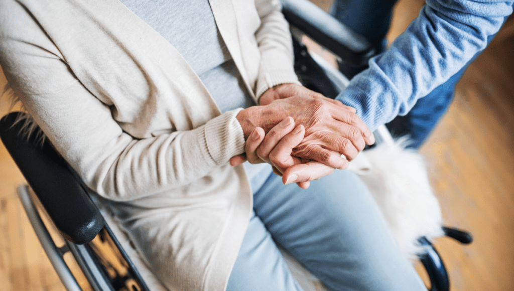 Plötzlich Pflegefall: Eine ältere Frau sitzt im Rollstuhl und hat ihre Hände um die einer zweiten Person gelegt, die sich zu ihr herunterzubeugen scheint.