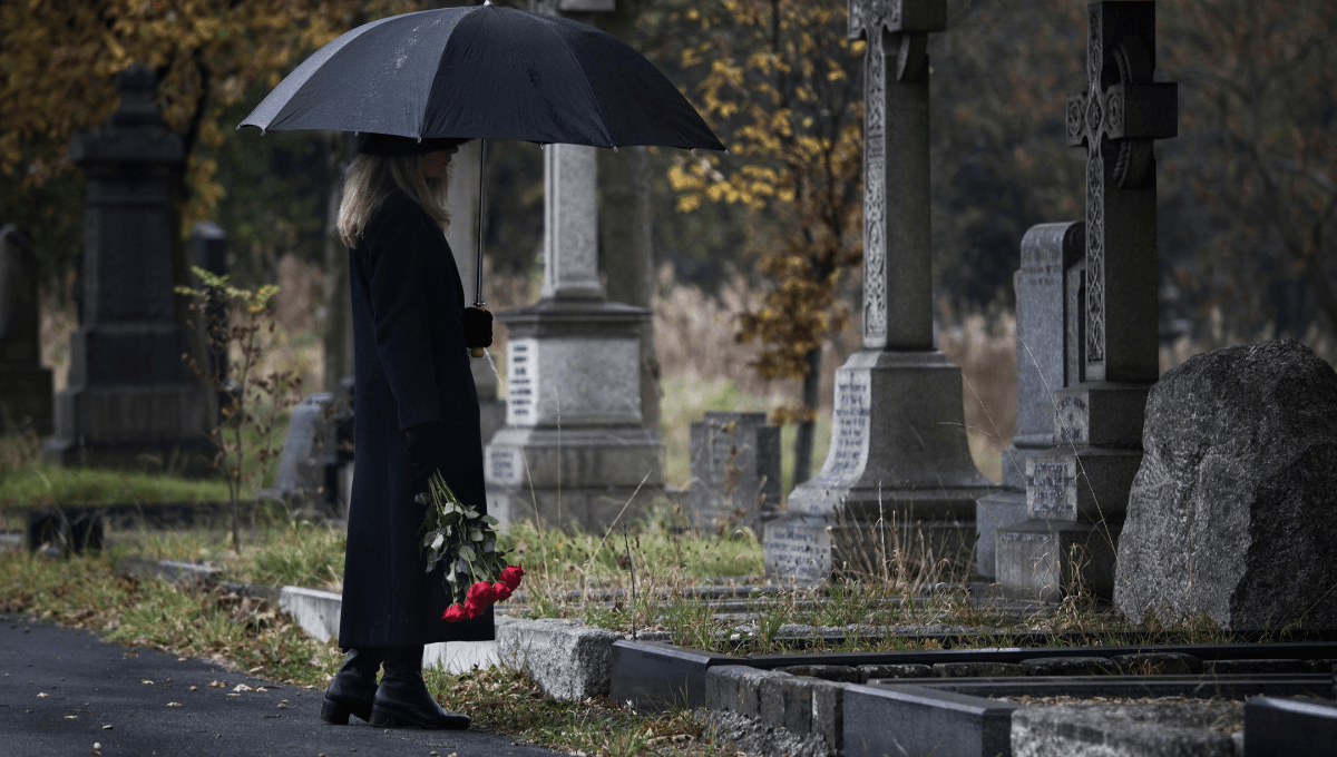 Eine Frau steht mit einem Strauß Blumen an einem Grab.