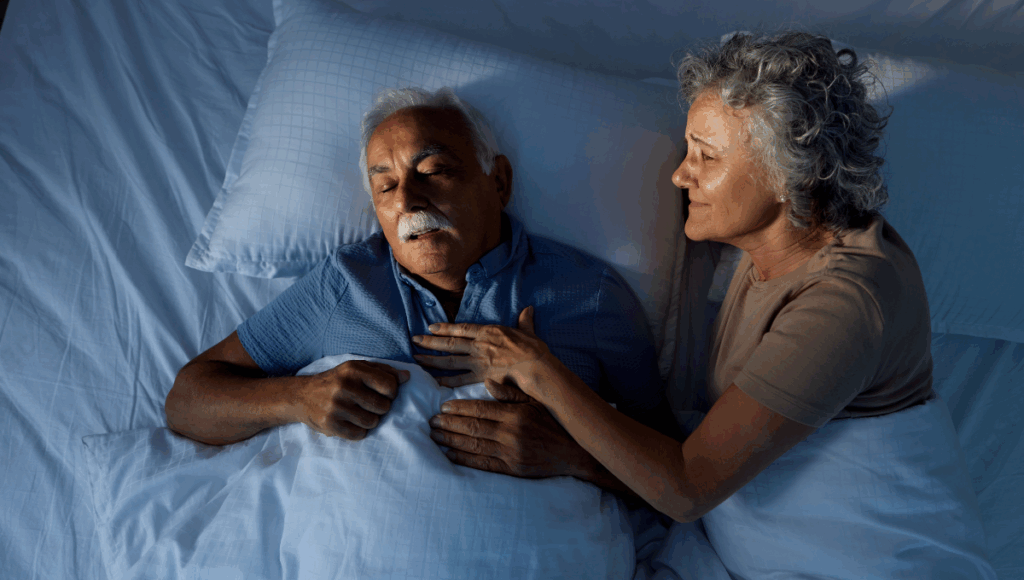 Ein Mann liegt nachts mit seiner Frau im Bett und zeigt Symptome für eine Schlafapnoe.