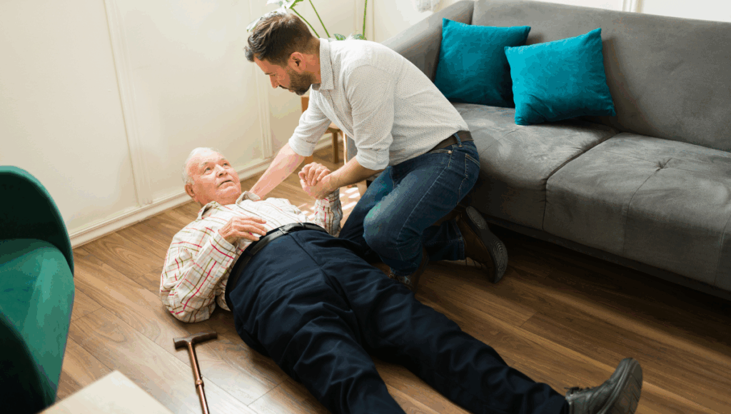Ein Senior erleidet einen Sturz und liegt auf dem Boden im Wohnzimmer. Sein Sohn leistet Erste Hilfe.